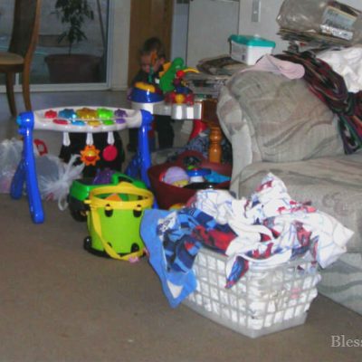 House cleaning does not need to be an overwhelming task. Just work some each day to get things done. Image of a room with a lot of toys, an overflowing laundry basket, and a couch cluttered with items. Blessed Catholic Mom watermark on photo at the bottom.