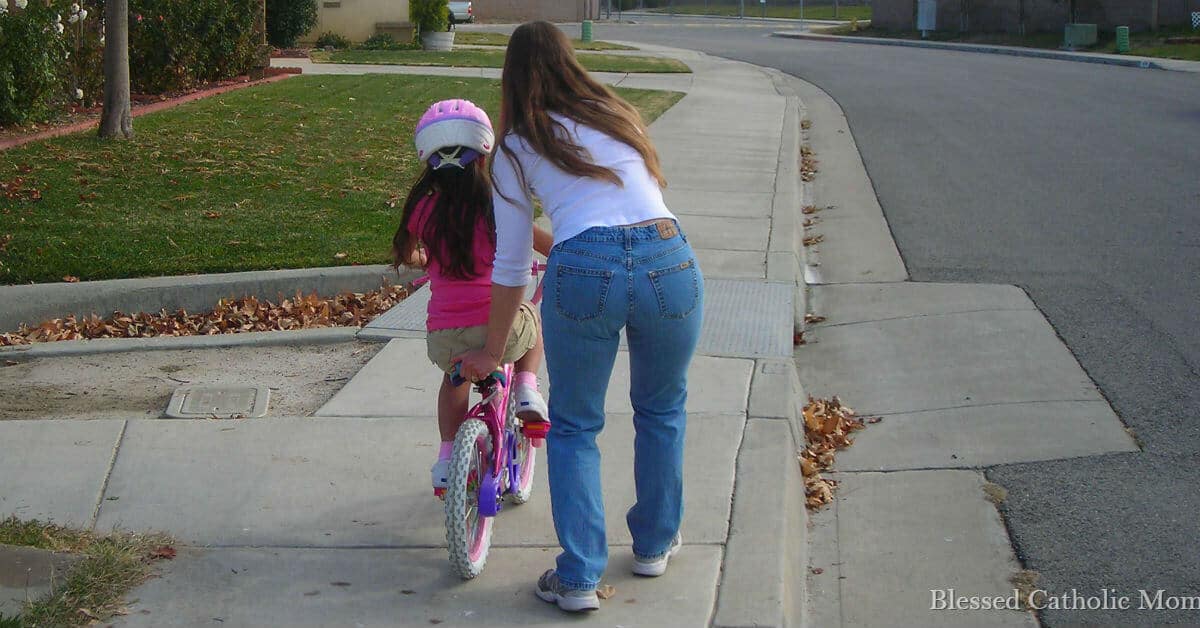 I can work each day as a mom to set a great example for our kids. Being a great mom does not equal being a perfect mom. I can rely on God's help. Image of a mom helping her daughter to learn to ride a bike without training wheels. Blessed Catholic Mom logo on image in right corner.