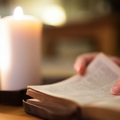 Image of a an open Bible with a hand about to turn the page. A lit candle in the background.