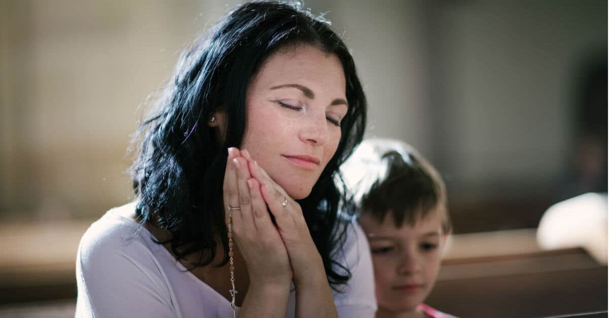 Image of a mom holding her Rosary and praying in church while her son sits beside her.