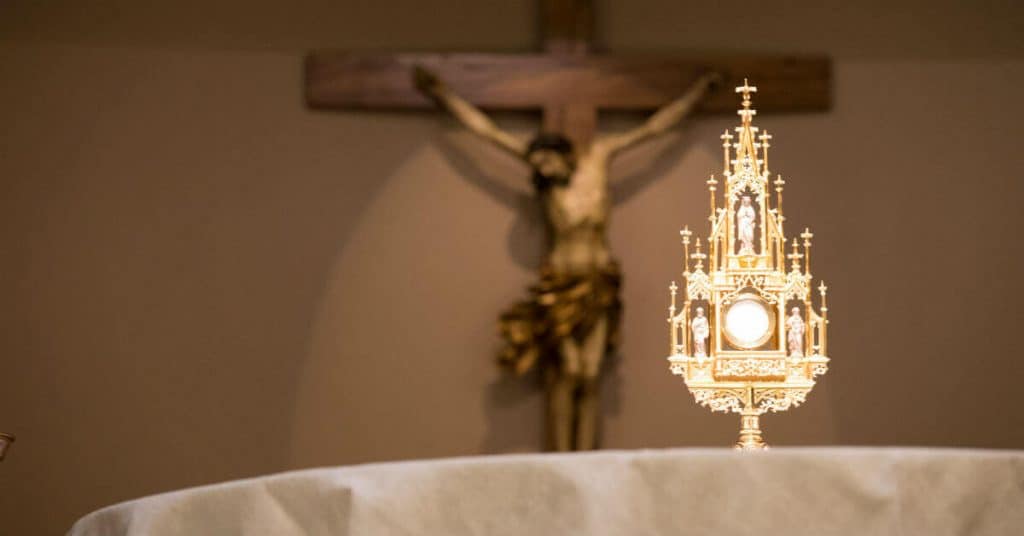 Image of an altar with a monstrance containing a host. In the background is a wooden crucifix: Jesus on the cross.