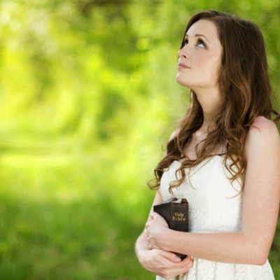 Image of a woman in a white dress holding a Bible, standing outside by trees, and looking up at the sky.
