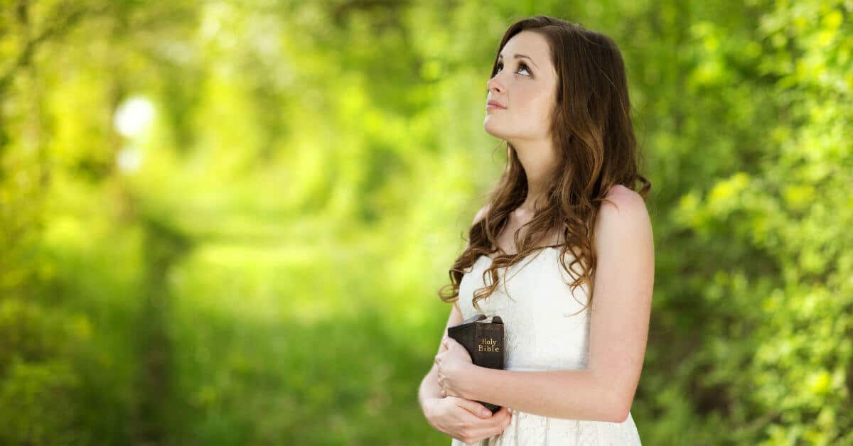 Image of a woman in a white dress holding a Bible, standing outside by trees, and looking up at the sky.