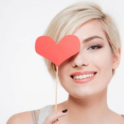 Image of a smiling woman with short blond hair holding a red heart on a stick in front of her right eye.