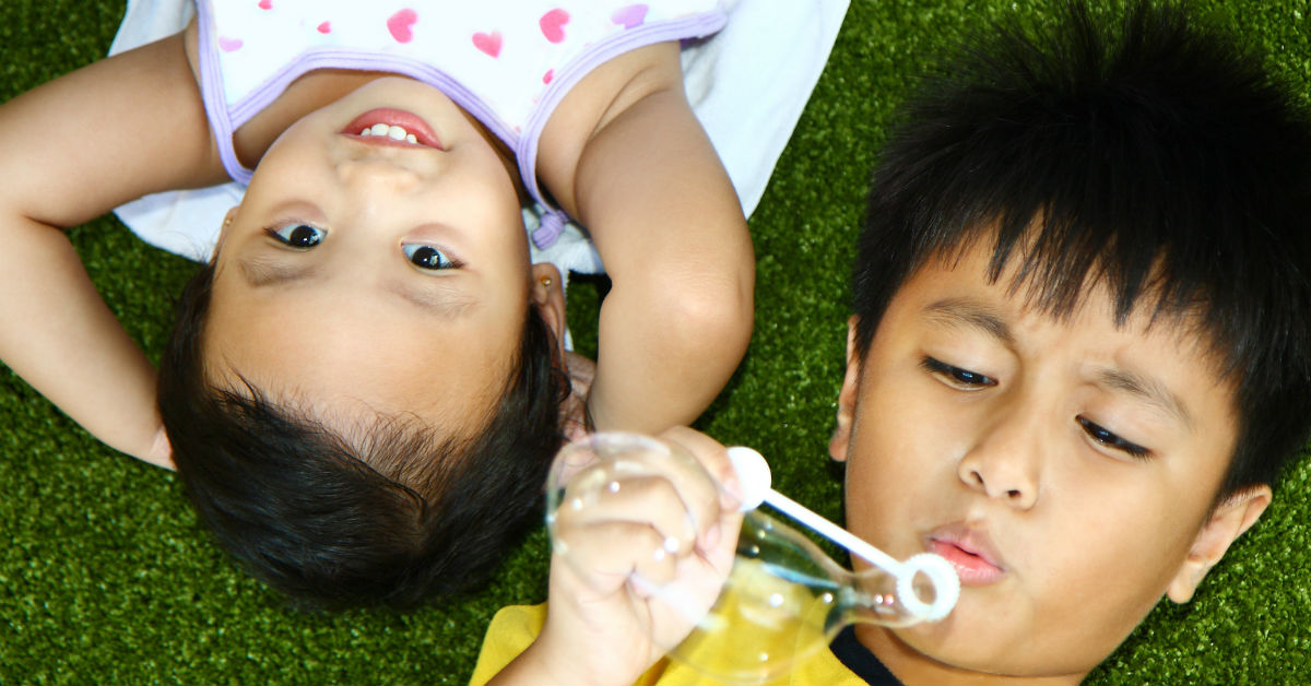 Image of a young boy and a young girl laying on their backs on the grass by each other. The boy is blowing soap bubbles.