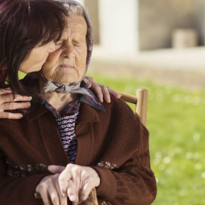 Image of a young woman leaning over to kiss on the cheek an older woman who is sitting down on a chair outside while she is holding a cane and has her eyes closed.