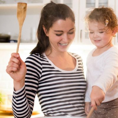 Image of a mom holding her daughter as the daughter is stirring food in a pot on the stove.