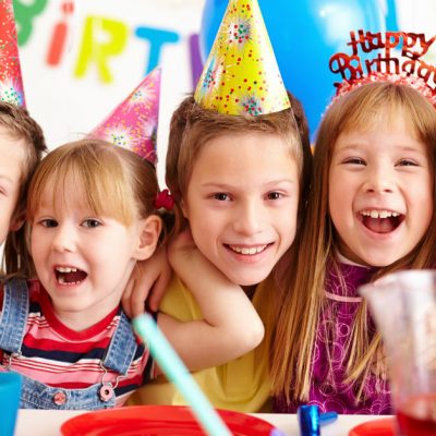 Image of five kids at a birthday party gathered together behind a table with food all smiling at the camera.