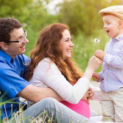Image of a man and his pregnant wife sitting together outside while their happy little son brings them a dandelion flower.