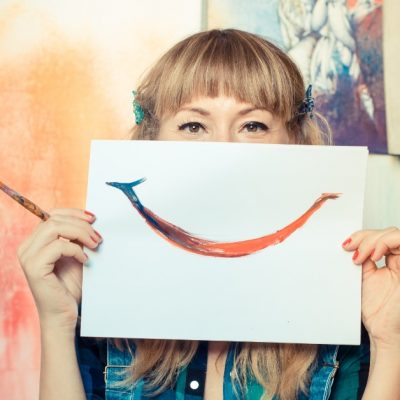 Image of a woman holding a paper in front of the lower half of her face that has a smile painted on it. She is holding a paint brush in one hand.