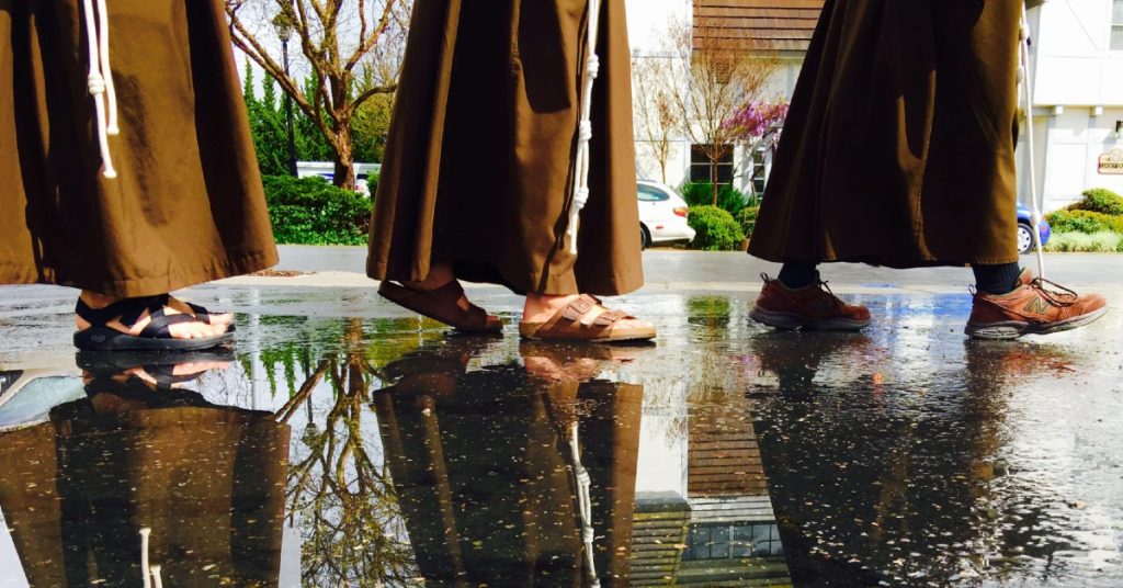 Partial image of three Franciscan men walking through a puddle of water with their reflections showing in the water beside them.