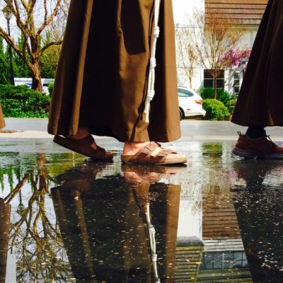 Partial image of three Franciscan men walking through a puddle of water with their reflections showing in the water beside them.