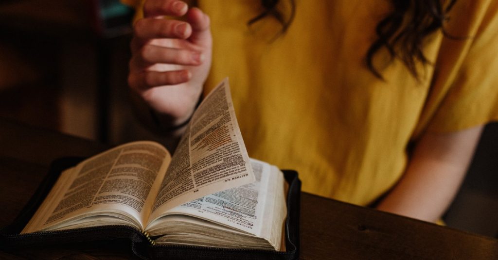 Image of a woman sitting at a table turning a page in the Bible that is open on the table in front of her.