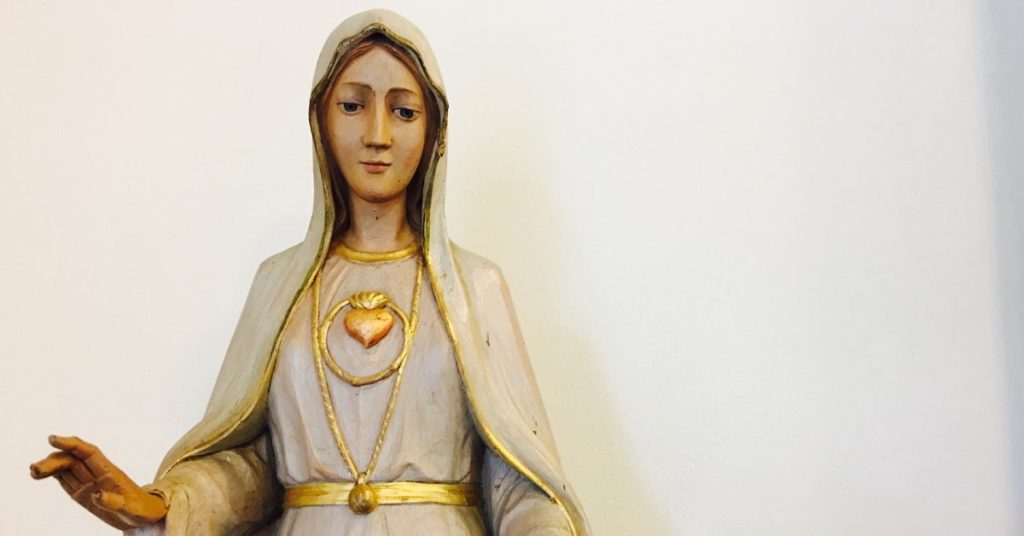 Image of a statue of the Blessed Virgin Mary. She is wearing white trimmed in gold and her arms are extended out toward whomever is in front of her. 