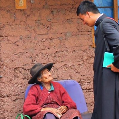 Image of a priest in his black habit wearing a backpack and holding a book under his arm, standing in front of an old woman who is sitting on a chair outside in front of an adobe looking brick home.