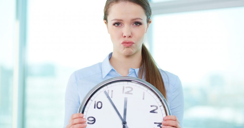 Image of a woman with a frustrated look on her face holding a large circular clock in front of her.