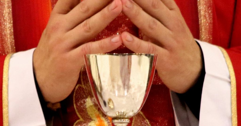 Image of a priest in red vestments with his hands together and fingers touching standing at the altar with a chalice in front of him.