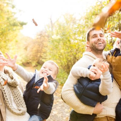 Image of a family (a mom and dad carrying their three boys) on a walk in colorful autumn forest and throwing leaves in the air.