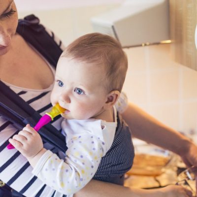 Image of a young mom with inner peace smiling down on her baby who she is wearing in a pack on her chest as she washes a glass in the kitchen sink.