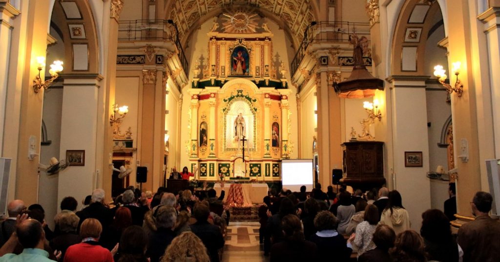 Image of the inside of a Catholic church during Mass. The camera is facing the altar and the pews are filled with people standing facing the altar.