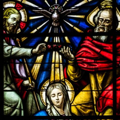 Image of a stained glass window showing the crowing of Mary as Queen of Heaven and earth by Jesus, the Holy Spirit, and God the Father.