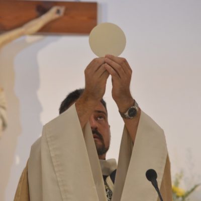 Image of a priest at the altar holding the consecrated host-the body of Christ-elevated for everyone to see and worship. Behind him is a representation of Jesus crucified.