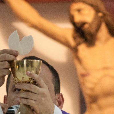 Image of a priest during the Catholic Mass holding up a broken Host above a Chalice. A wooden image of Jesus crucified is on the wall behind him.