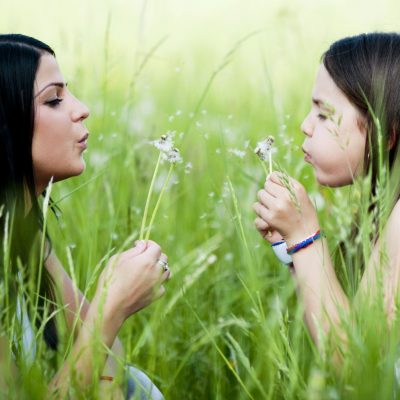 Image of a woman and her daughter sitting in a field facing each other and each of them are blowing a dandelion.