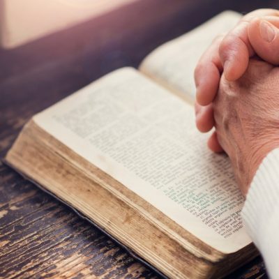 Image of a Bible open on a table by a window and the hands of a woman are clasped together on top of the Bible.