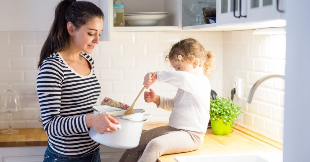 Image of a mom holding a pot of something out to her little daughter who is sitting on the counter and mixing it.
