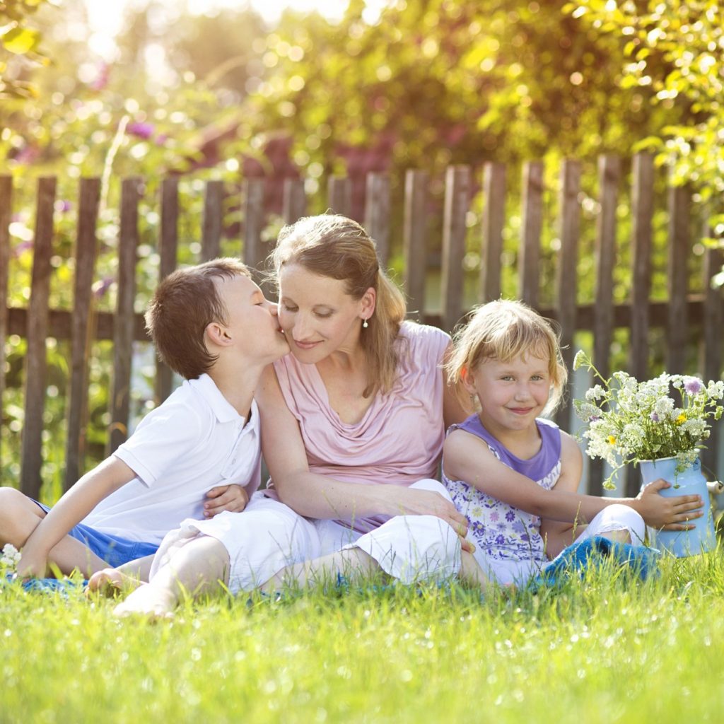 Image of a happy mother with her two children-one boy giving her a kiss and her daughter- spending time together sitting outside in nature.
