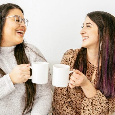 Image of two women sitting on a couch beside each other, smiling and sharing coffee together.