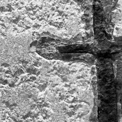 Image in black and white of a concrete wall with a cross etched in the wall that is darker than the wall.