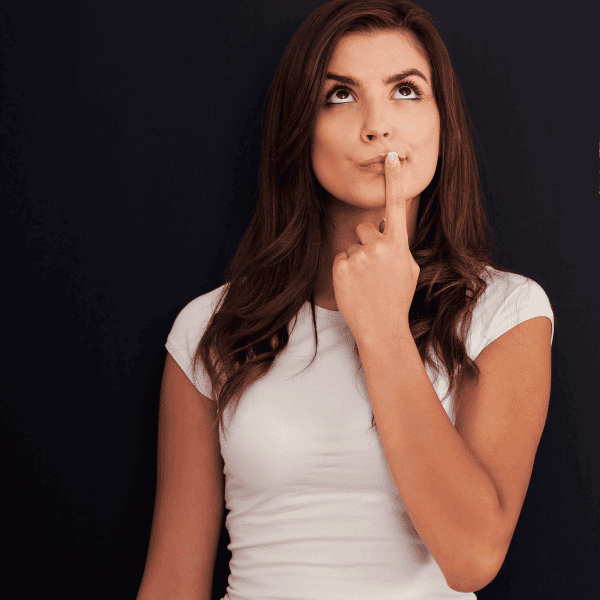  Image of a woman in a white t-shirt standing with her left index finger against her closed mouth, looking up.